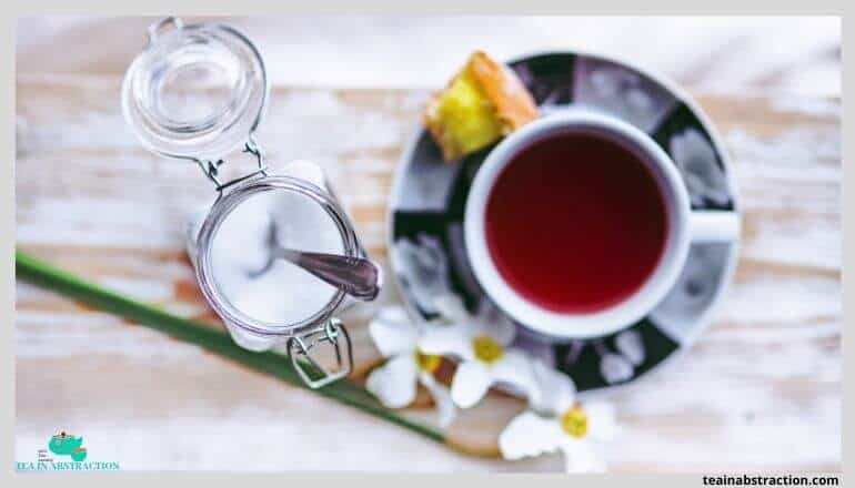 red hibiscus tea in a white cup on a saucer sitting oon a table next to an open jar of sugar with a spoon in it. flowers adorn the image as well