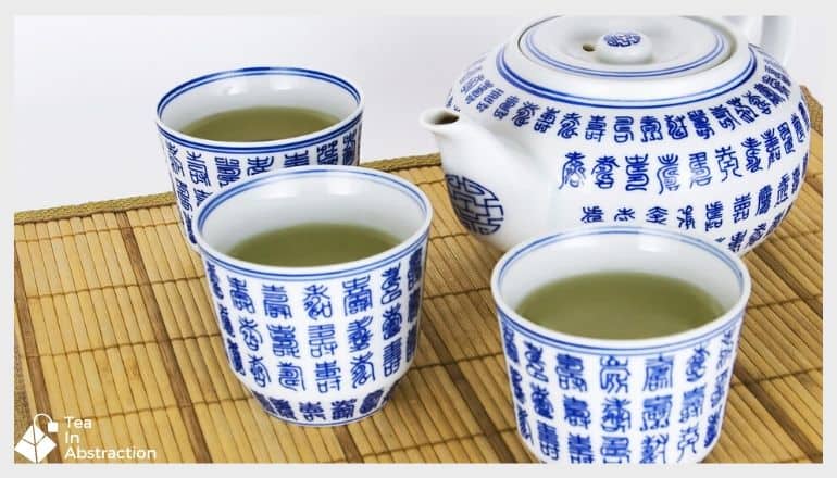 3 cups of green tea in decorative tea cups next to a tea kettle. white with blue writing on them